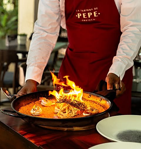 Arroz en paellera al fuego, con camarones y llamas, sostenido por un cocinero con delantal de la casa
